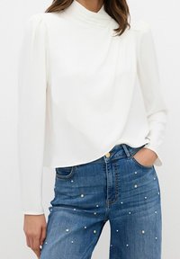 Femme portant un chemisier blanc à manches longues avec un col plissé et un jean bleu orné de petites perles décoratives.