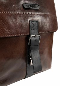 Brązowa skórzana torba z czarnym paskiem, z metalową klamrą i fakturowanym wykończeniem. Klapa prezentuje wyciśnięte logo u góry.