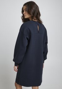Navyblaues, übergroßes Sweatshirt-Kleid mit langen Ärmeln, einem Schlüsselritzenausschnitt auf der Rückseite und Seiten-Schlitzen am Saum. Glatte Stofftextur.