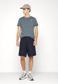 Tricou gri, pantaloni scurți bleumarin și o șapcă bej deschis. Pantofi sport casual cu accente gri, roșii și albe. Design simplu, potrivire confortabilă.