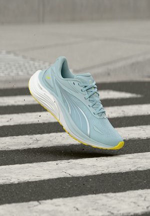 Chaussure de course bleu clair avec semelle jaune en l'air au-dessus d'un passage piéton rayé noir et blanc, petits gravillons volant autour.