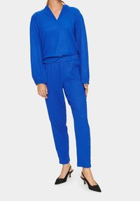 Modrý, lesklý dvoudílný outfit obsahující dlouhý rukáv s véčkovým výstřihem a kalhoty s elastickým pasem, doplněný černými lodičkami s špičatou špičkou.