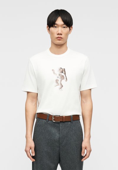 Homme portant un T-shirt blanc avec un graphisme d'astronaute, un pantalon gris et une ceinture marron, debout devant un fond gris uni.