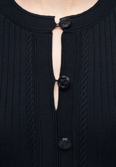 Κοντινό πλάνο σε μαύρο ριμπ πλεκτό τοπ με τρία διακοσμητικά μαύρα κουμπιά, δύο κουμπωμένα και ένα ξεκουμπωμένο στο λαιμό και το στήθος.