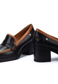 Mocasines de cuero negro con acentos marrones y una correa cosida, con un tacón cuadrado y detalles en hardware dorado.