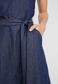 Džínové šaty v tmavě modré barvě, s uvázaným páskem v pase, bez rukávů a bočními kapsami. Materiál má hladký, strukturovaný povrch.