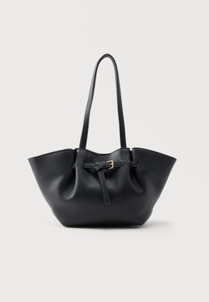 Zwarte leren tote bag met lange handvatten en een voorband met een kleine gouden gesp, tegen een effen achtergrond.