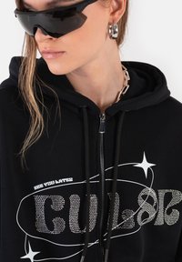 Czarna bluza z kapturem zapinana na zamek błyskawiczny z tekstem „CULT” i „SEE YOU LATER” wykonanym z srebrnych kryształków. Model nosi czarne okulary przeciwsłoneczne i łańcuchowy naszyjnik.