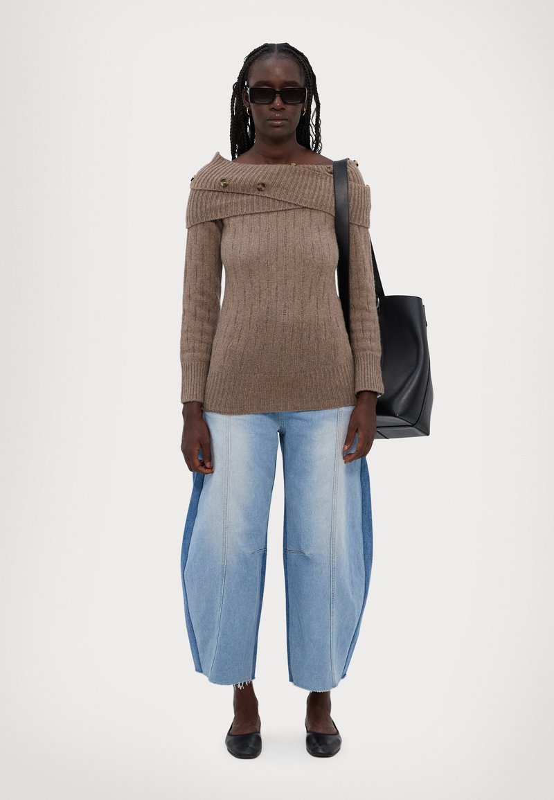Brauner, gerippter Pullover mit schulterfreiem Design und zwei Knöpfen; kombiniert mit weiten, hellblauen Jeans mit Seiteneinsätzen; schwarze Tragetasche.