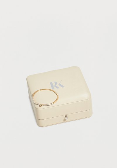 Μπεζ τετράγωνο κουτί κοσμημάτων με ασημένιο λογότυπο "RK", κλειστό, με λεπτό χρυσό βραχιόλι να ακουμπά πάνω του.