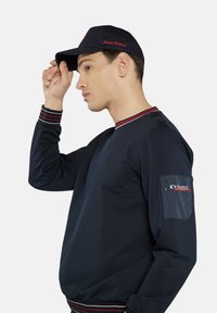 Granatowy sweatshirt z czerwonymi paskami przy mankietach i dekolcie. Posiada kieszeń naszywaną z logo "ICY FLANKS". Nosi się z dopasowaną czapką.