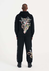 Schwarzer Hoodie mit buntem Grafikdesign auf der Rückseite, passende Jogginghosen mit seitlichem Text. Das Material scheint weich zu sein; beide haben Bündchen.