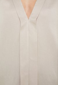 Vêtement beige à col en V avec une couture centrale verticale à l'avant.