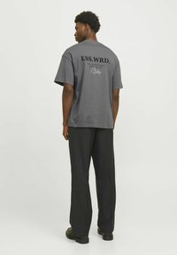 Grå bomulds-T-shirt med "ESS.WRD." trykt i sort på ryggen, parret med løse sorte bukser og sorte sko.