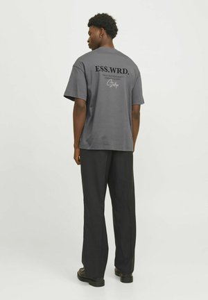 Grå bomull t-shirt med "ESS.WRD." tryckt i svart på baksidan, tillsammans med lösa svarta byxor och svarta skor.