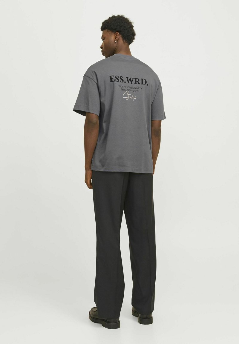 Grå bomulds-T-shirt med "ESS.WRD." trykt i sort på ryggen, parret med løse sorte bukser og sorte sko.