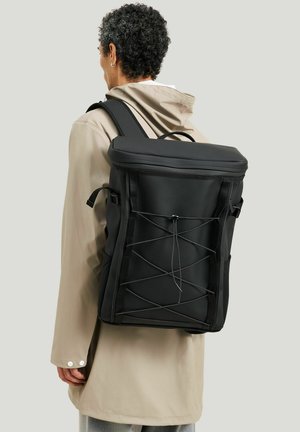 Person iført en beige hættetrøje, der bærer en stor sort rektangulær rygsæk med elastisk snordetalje foran, set bagfra.