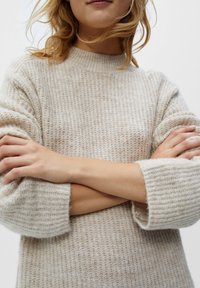 Pull en tricot beige avec une texture côtelée, col montant et manches surdimensionnées, présentant une apparence douce et chaude.