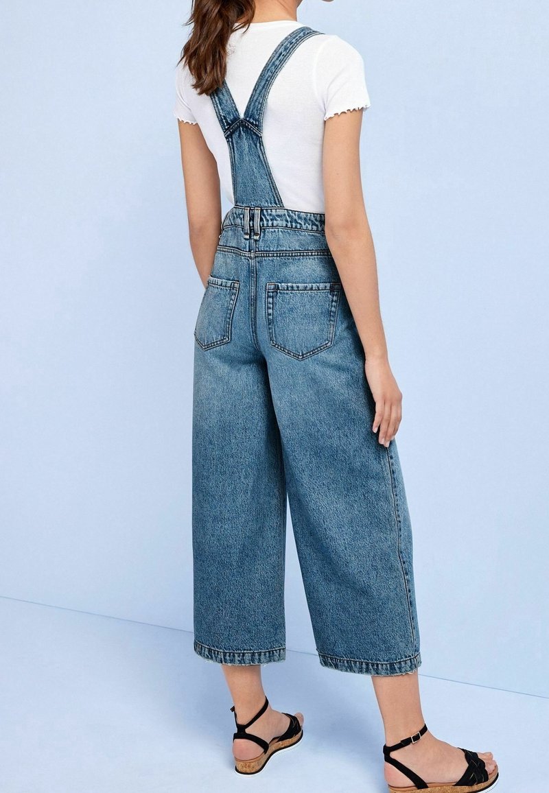 Helle blaue Denim-Latzhose mit weit geschnittenen Beinen, verstellbaren Trägern und Gesäßtaschen, kombiniert mit einem weißen, taillierten Shirt und schwarzen Sandalen.