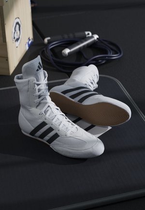 Witte Adidas boksschoenen met zwarte strepen op een zwart matje, naast een opgerolde zwarte springtouw en een houten doos.
