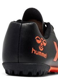 Scarpa da calcio sintetica nera con superficie texturizzata, dettagli arancioni e un prominente logo 'hummel', caratterizzata da una suola nera robusta per la trazione.