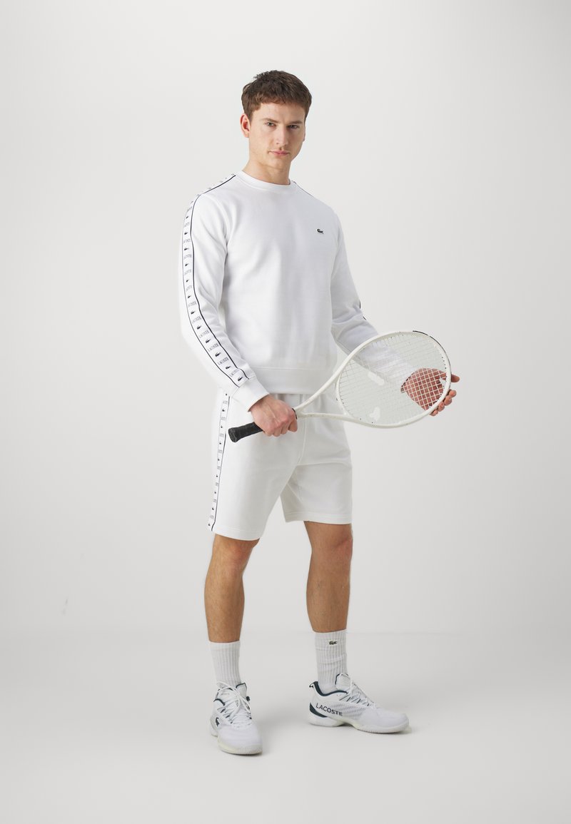 Felpa atletica bianca e pantaloni shorts con accenti neri sui lati, abbinati a sneakers bianche e una racchetta da tennis. Design pulito e minimalista.