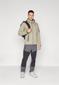 Light beige zip-up jacket with hood, dark gray and black pants with pockets, worn with a black backpack and gray sneakers.