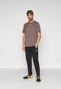 T-shirt de algodão castanha com um logótipo amarelo, combinada com calças pretas com um gráfico branco. Calçado preto com detalhes em bege.