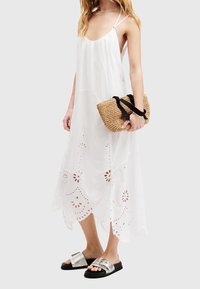 Femme portant une robe midi blanche sans manches avec des découpes florales, tenant un sac en paille avec des anses noires, et des sandales argentées à enfiler.