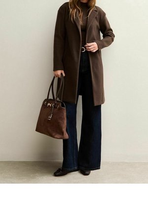 Vrouw met een bruine jas, donkerdere wijde jeans, bruine platte schoenen, met een grote bruine leren handtas tegen een effen lichte muur.