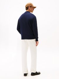 Maglione navy dal taglio rilassato, abbinato a pantaloni bianchi e scarpe nere. Il modello indossa un berretto marrone chiaro ed è posizionato di profilo.