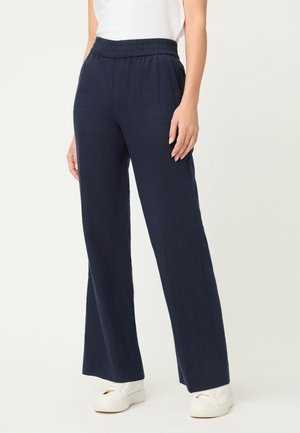 Donna che indossa pantaloni larghi blu navy con vita elastica e sneakers bianche, in piedi davanti a uno sfondo chiaro e uniforme.