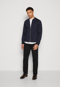 Marynarska nepremočljiva jakna z dvostranskim zapiranjem in stoječim ovratnikom, dolgi rokavi z rebrastimi manšetami, v kombinaciji z belo srajco s ovratnikom in črnimi hlačami.