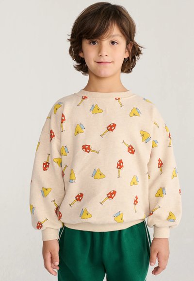 Enfant aux cheveux bruns courts porte un sweat crème avec des motifs de champignons rouges et jaunes et un pantalon vert, debout devant un fond uni.