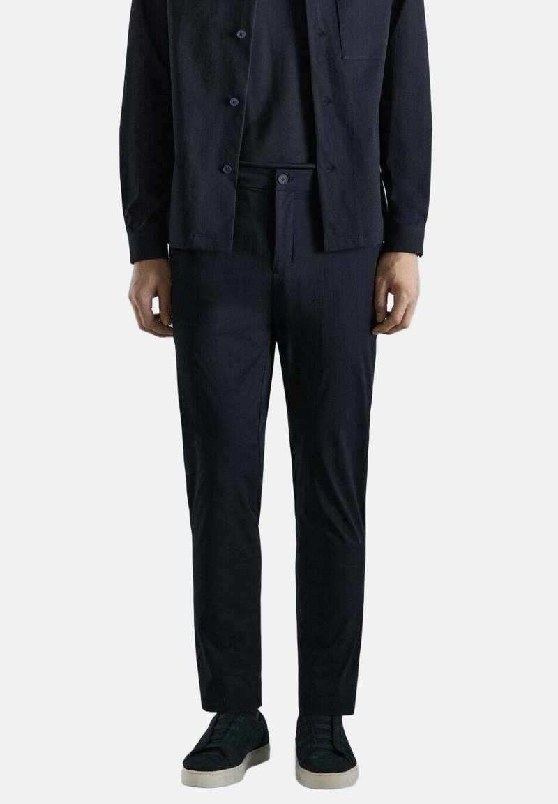 Homme debout portant une chemise à boutons noire, un pantalon noir et des baskets noires avec des semelles blanches, sur un fond clair uni.