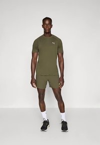 Camiseta deportiva y pantalones cortos de color verde oliva, diseño ajustado, material que absorbe la humedad, con logo en la camiseta, combinados con zapatillas negras y calcetines blancos.