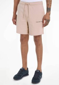 Pantalones cortos de algodón beige con cintura elástica y cordón; presenta el logo negro de "Calvin Klein Jeans" en el lado izquierdo.
