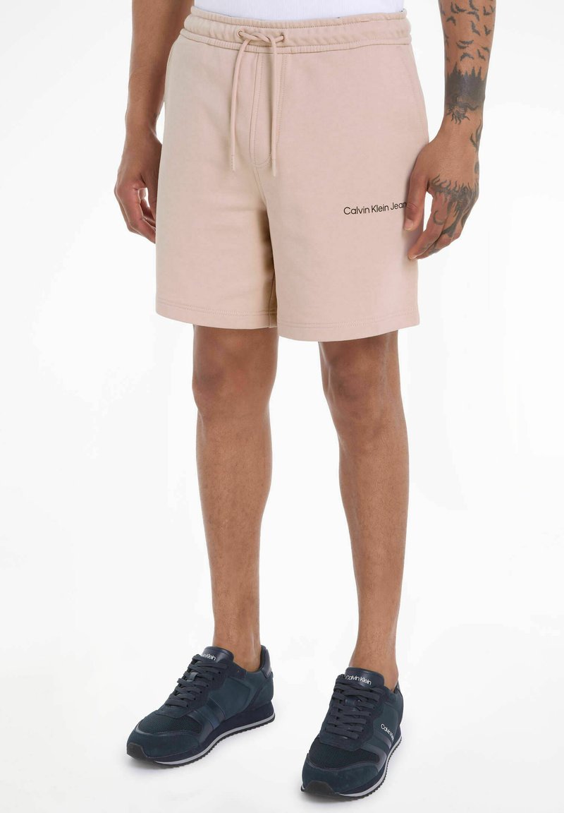 Pantalones cortos de algodón beige con cintura elástica y cordón; presenta el logo negro de "Calvin Klein Jeans" en el lado izquierdo.
