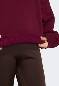 Bordový svetr s jemnou texturou, s ribkovaným lemem. Hnědé legíny s hladkým povrchem. Ruka držící spodní okraj svetru.