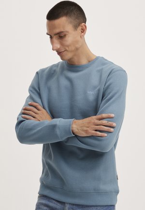 Light blue sweatshirt, crew neck, ribbed cuffs and hem, soft cotton blend material. Subtle logo on the chest, model wears arms crossed.