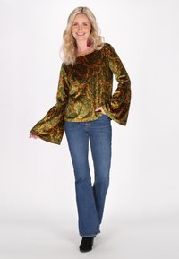 Top in velluto con motivo paisley verde e oro, con maniche a campana, abbinato a jeans blu a campana e stivaletti neri.