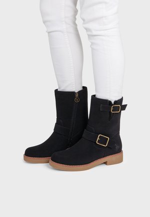 Bottes noires mi-mollet avec boucles latérales et fermetures éclair portées avec un pantalon blanc ajusté sur un fond clair.