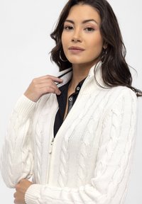 Kabelstrik, cremefarvet cardigan med høj krave og lynlås. Ribbet manchetter og kant. Bæres over en sort skjorte med knapper.