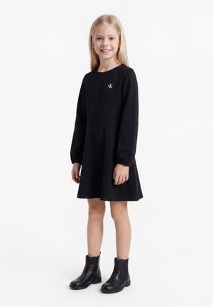 Ung pige med langt blondt hår iført en sort langærmet kjole og sorte ankelstøvler, stående og smilende mod en hvid baggrund.