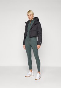 Veste courte en doudoune noire avec capuche, associée à un ensemble de leggings texturé vert foncé. Baskets blanches avec semelles marron clair.