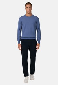 Mėlynas megztinis su apykakle, su ilgomis rankovėmis, derinamas su tamsiomis kelnėmis ir baltais sportbačiais, stovintis prieš paprastą foną.