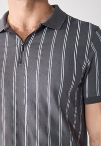 Szürke polo ing fehér függőleges csíkokkal, cipzáras gallérral, rövid ujjú és texturált anyag jellemzi.