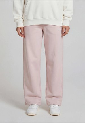 Person iført lyserosa vidåpne joggebukser og hvite joggesko, stående mot en ensfarget lys grå bakgrunn.