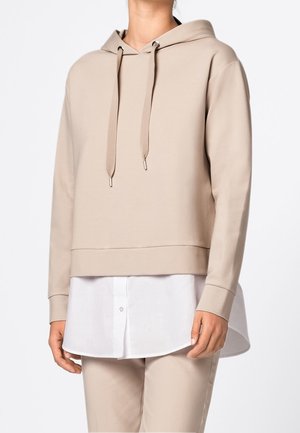 Persoon met een beige hoodie over een witte blouse met knopen, gecombineerd met bijpassende beige broek, tegen een effen lichte achtergrond.