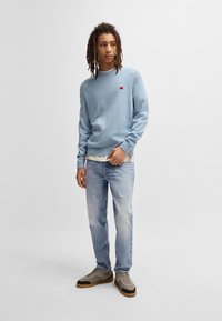 Sudadera de color azul claro con puños y dobladillo acanalados; pequeño logo rojo en el pecho. Combinada con jeans azul desgastados y zapatillas gris-marrón.